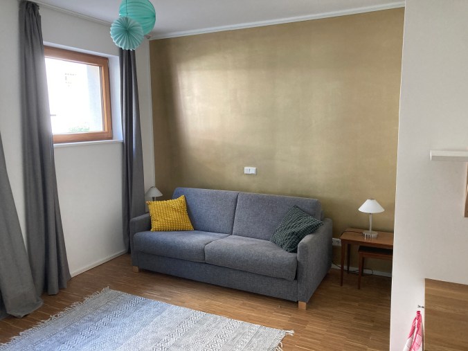 Ein Blick in unser Gästezimmer mit Teppich im Vordergrund, dahinter ein (zusammengeklapptes) Schlafsofa und dahinter eine goldfarbene Wand