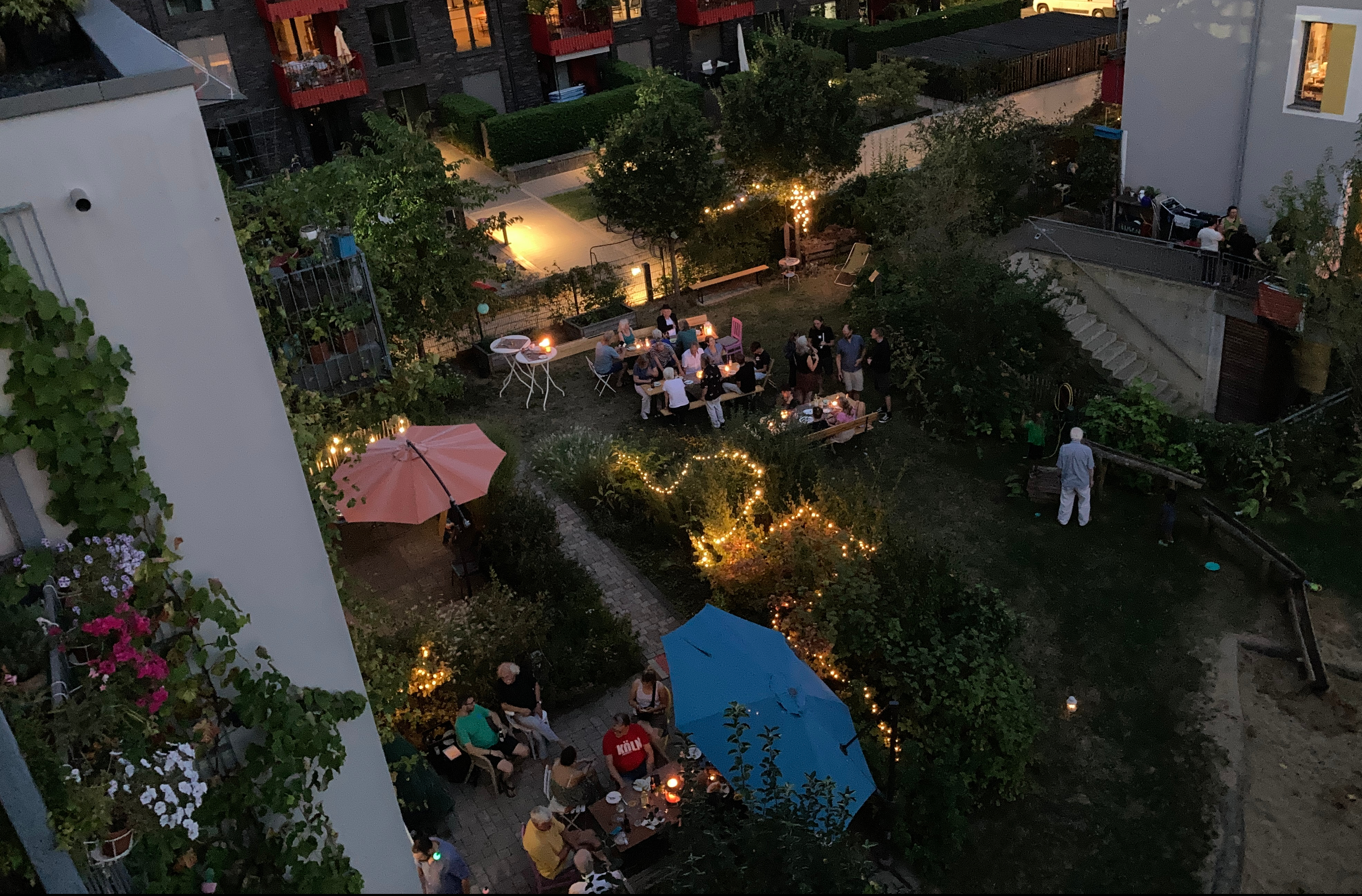 Bild eines Gartens in der Abend-Dämmerung von schräg oben. Man erkennt einige Menschen um Tische sitzend. An verschiedenen Stellen leuchen Lichterketten.