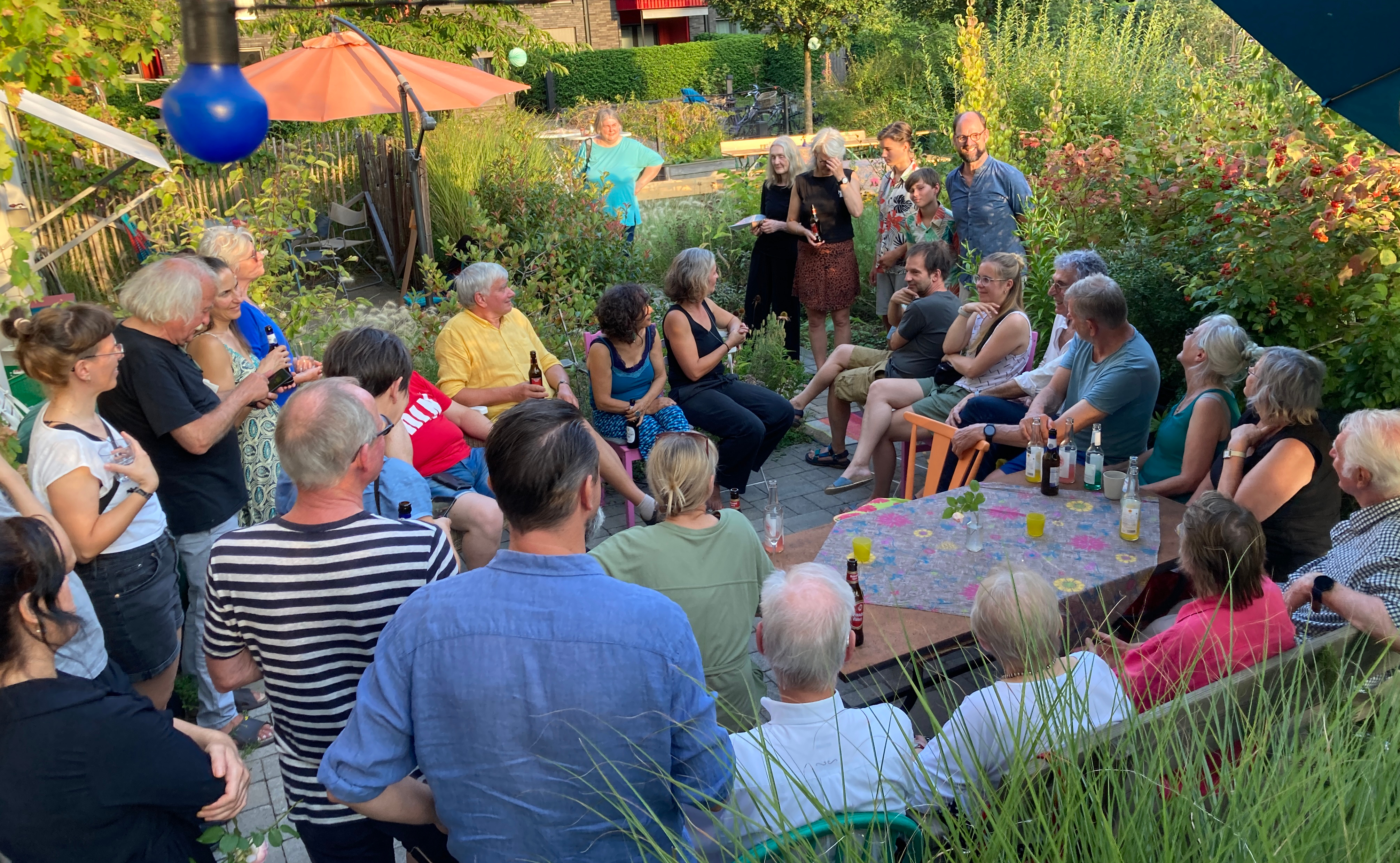 Ca. 30 Personen sitzen bzw. stehen im Kreis bei sonnigem Wetter in einem attraktiven Garten